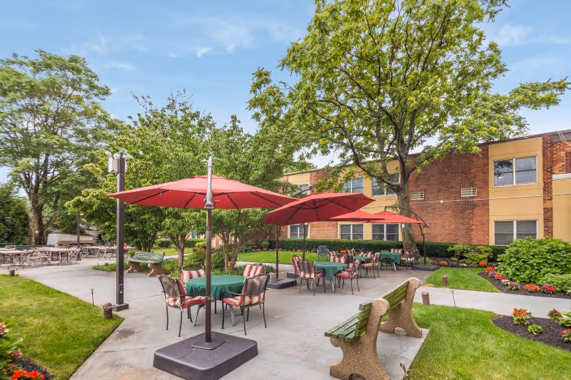 Outdoor patio area at Castle Senior Living with several tables covered with green tablecloths and red umbrellas. There are cushioned chairs around the tables, green grass, trees, and a building with brick and tan siding in the background.