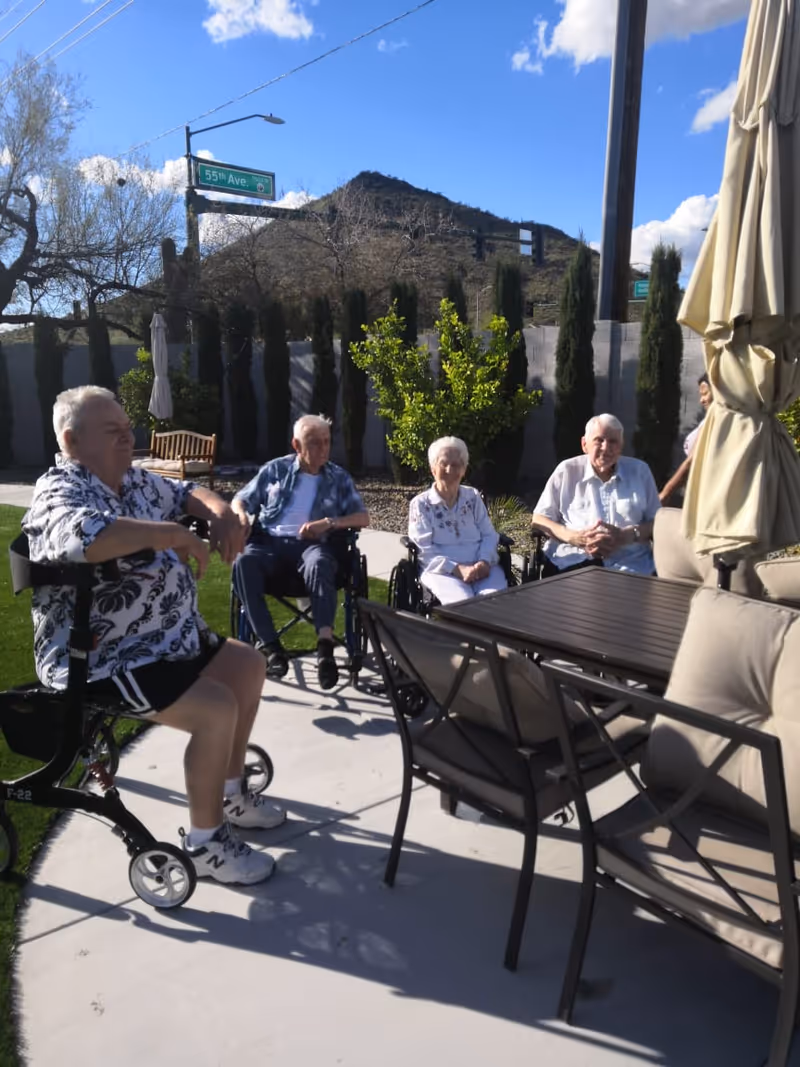 Four elderly individuals sitting outdoors on a sunny day, three in wheelchairs and one on a walker with a seat, gathered around a patio table with chairs and an umbrella. Behind them are tall trees, a concrete wall, and a mountain in the background under a blue sky with some clouds.