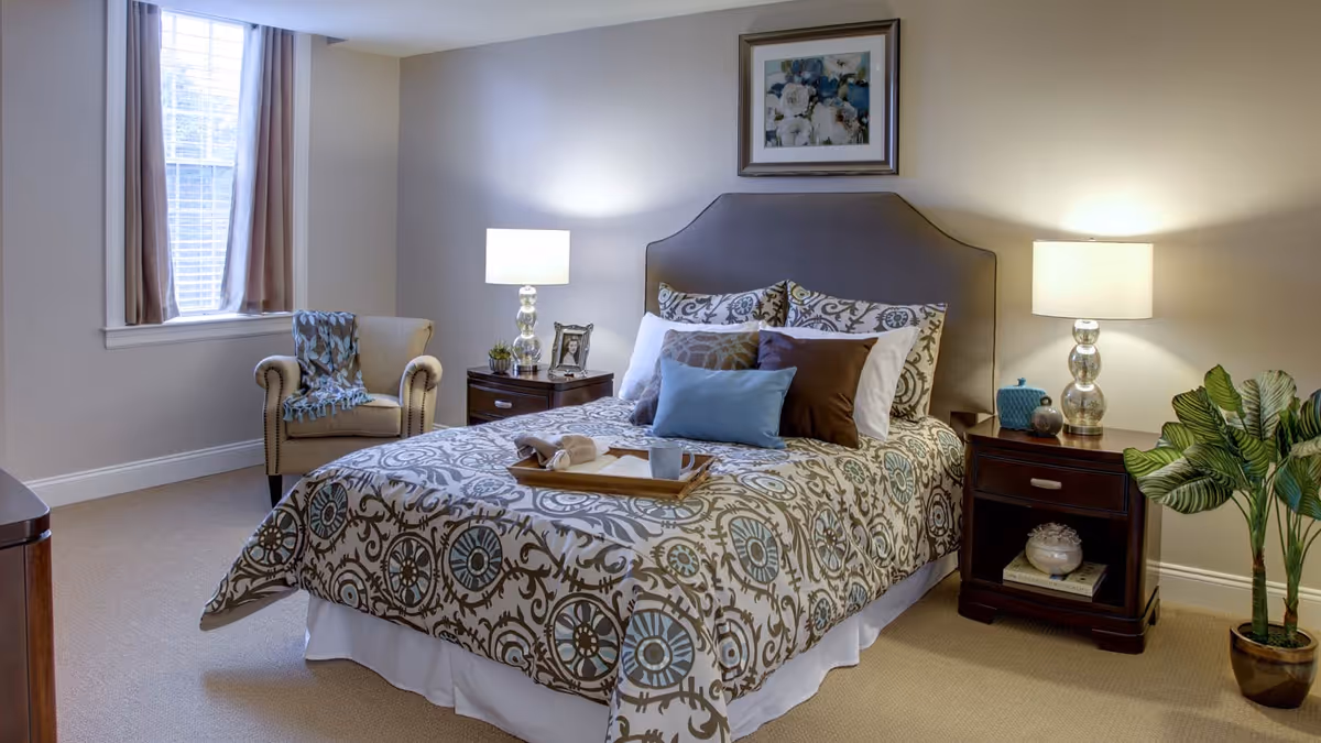 A cozy bedroom with a patterned bedspread on a double bed, multiple pillows, two bedside tables with lamps, a framed floral picture on the wall, a beige armchair with a throw blanket near a window with curtains, and a potted plant on the floor.