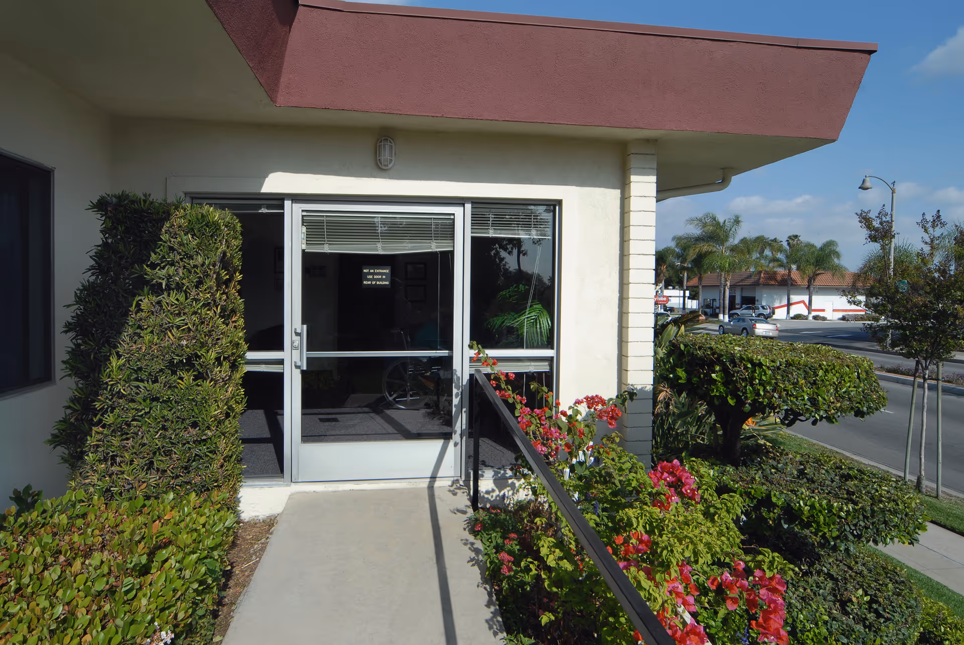 Entrance to La Habra Convalescent Hospital showing a glass door with a sign, surrounded by trimmed bushes and flowering plants along a concrete walkway, with a street and palm trees visible in the background under a clear sky.