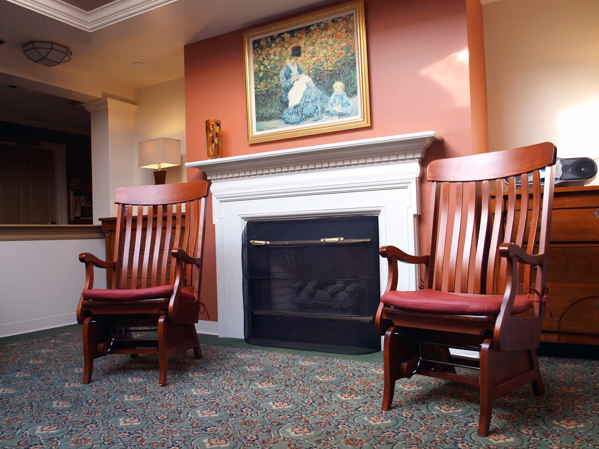 A cozy interior room with two wooden armchairs with red cushions placed on a patterned carpet in front of a white fireplace. Above the fireplace is a framed painting of a woman and child in a garden. A table lamp and a decorative vase are on the mantel, and a wooden sideboard with a speaker is visible to the right.