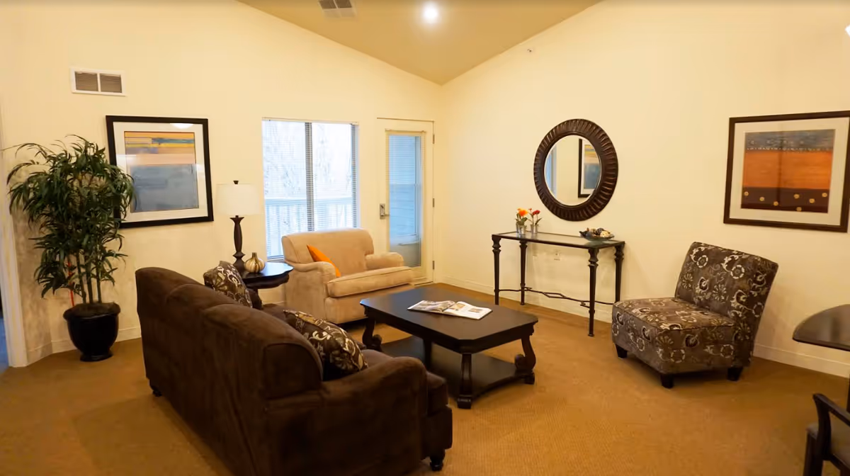 A cozy living room with a brown sofa, a beige armchair, a patterned chair, a dark wooden coffee table with an open book, a side table with a lamp, a console table with decorative items and a round mirror above it, two framed artworks on the walls, a potted plant in the corner, and a door with a window leading outside.