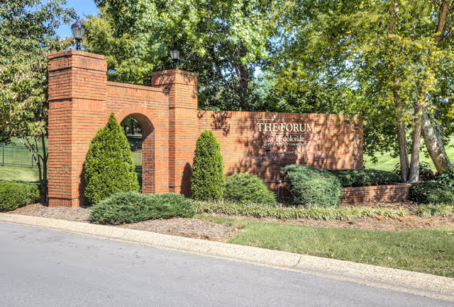 Brick entrance sign for The Forum at Brookside surrounded by green shrubs and trees, with a paved road in the foreground.