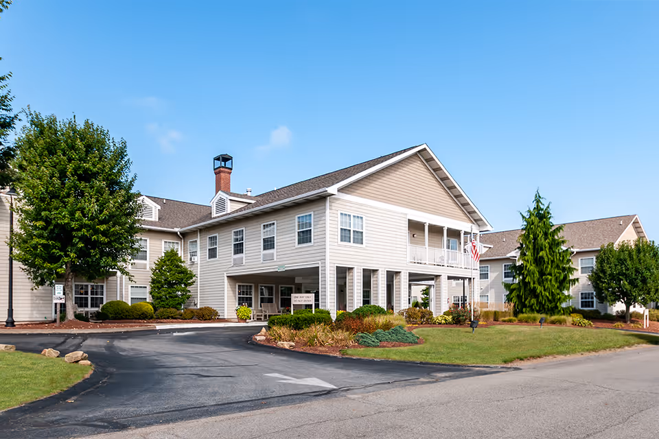 Exterior view of a two-story senior living facility building with beige siding, white trim, and a covered entrance. The building is surrounded by green lawns, trees, and shrubs under a clear blue sky.