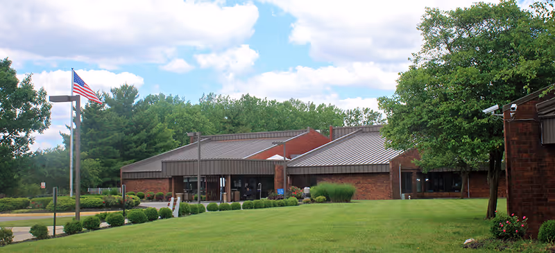 Exterior view of a single-story brick building with a metal roof, surrounded by green grass, bushes, and trees under a partly cloudy sky. An American flag is flying on a flagpole near the entrance.