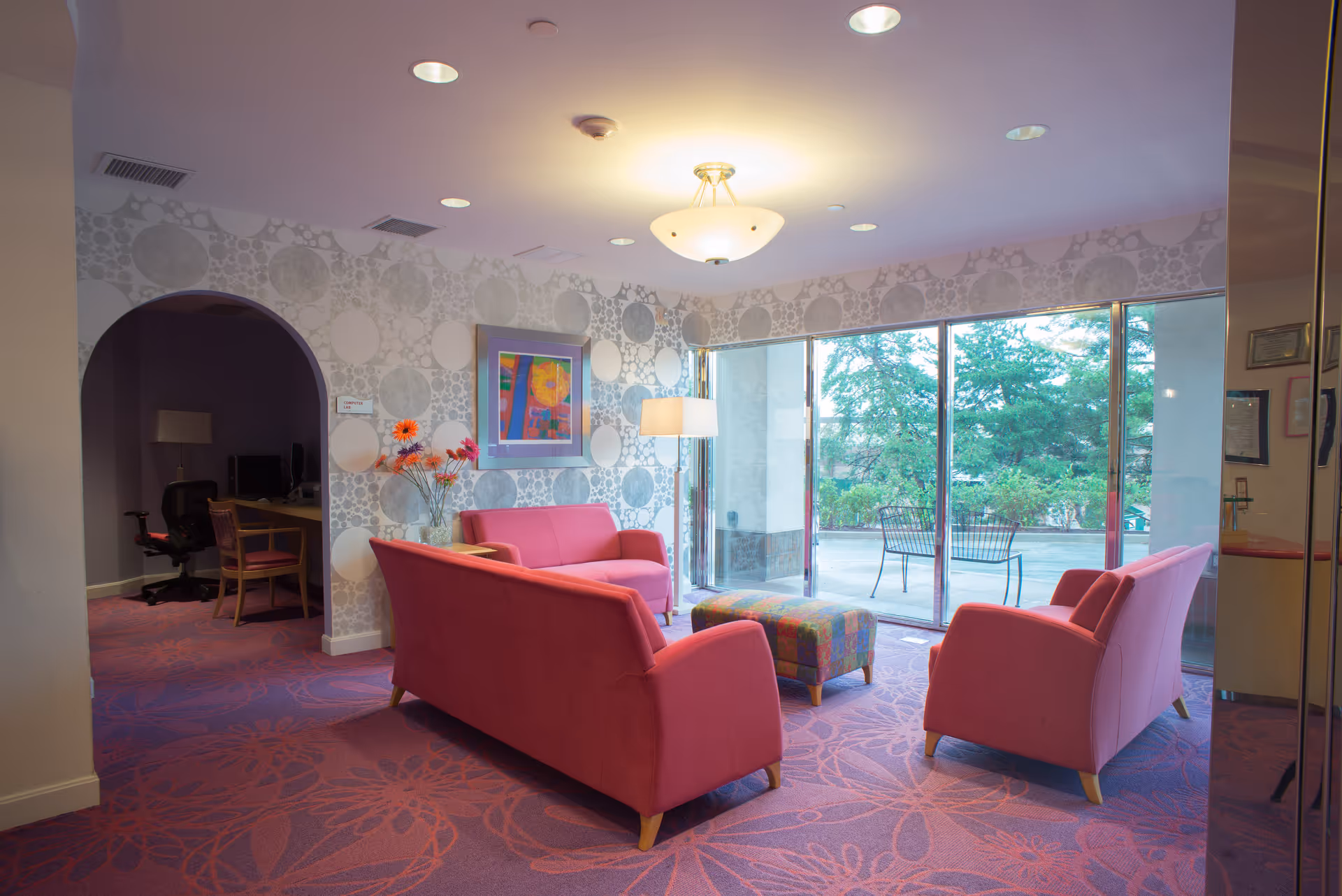 A cozy living room area with pink upholstered sofas and a colorful ottoman on a purple carpet with floral patterns. There is a floor lamp beside one sofa, a vase with orange and pink flowers on a small table, and a framed abstract painting on the wall. The room has large sliding glass doors that open to an outdoor patio with metal chairs and greenery visible outside. To the left, there is an arched doorway leading to a computer lab with desks and chairs.