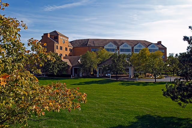 Exterior view of a large brick building with multiple windows and a sloped roof, surrounded by green trees and a well-maintained lawn under a clear blue sky.