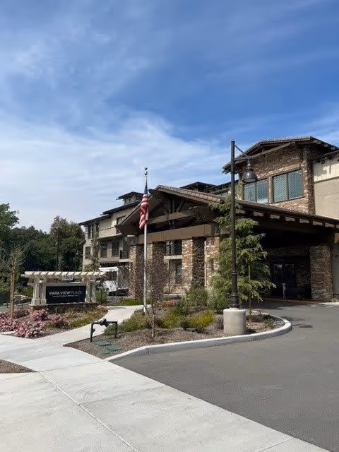 Exterior view of Park View Place Senior Living facility showing a multi-story building with stone and wood accents, an American flag on a flagpole, landscaped greenery, and a paved driveway under a partly cloudy sky.