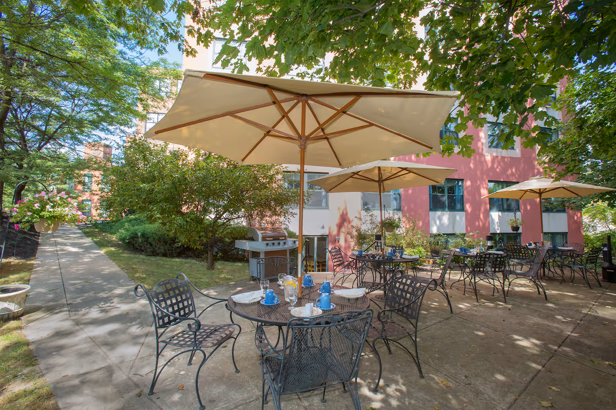 Outdoor patio area with several round metal tables and chairs, each table shaded by large beige umbrellas. The tables are set with blue cups, plates, and pitchers. The patio is surrounded by greenery including trees and bushes, with a pink building in the background.