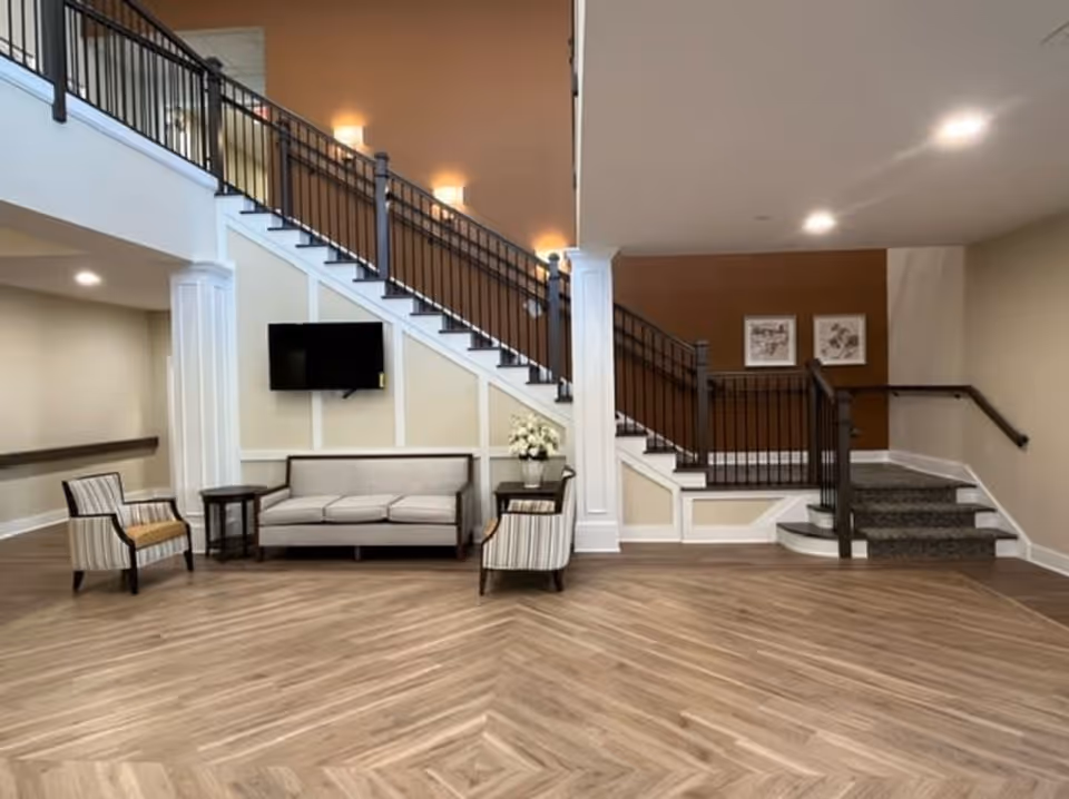 A spacious interior common area featuring a staircase with black railings and beige walls. Below the staircase is a seating area with a beige sofa, two striped armchairs, and two small wooden side tables. A flat-screen TV is mounted on the wall above the sofa, and a vase with white flowers is placed on one of the side tables. The floor has a wood-patterned design, and the lighting is warm and inviting.