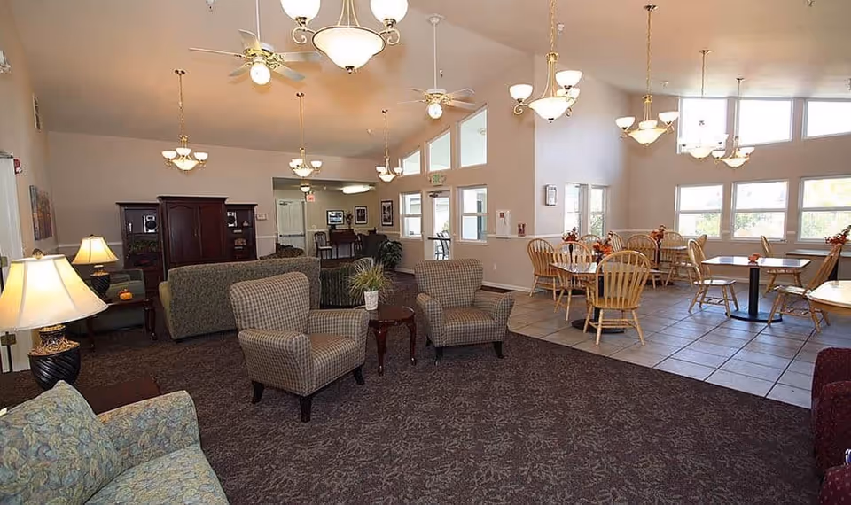 A spacious senior living common area with a mix of seating including armchairs and sofas on carpeted flooring, and a dining area with wooden tables and chairs on tiled flooring. The room has high ceilings with multiple ceiling fans and hanging light fixtures, large windows letting in natural light, and a few decorative plants and lamps.