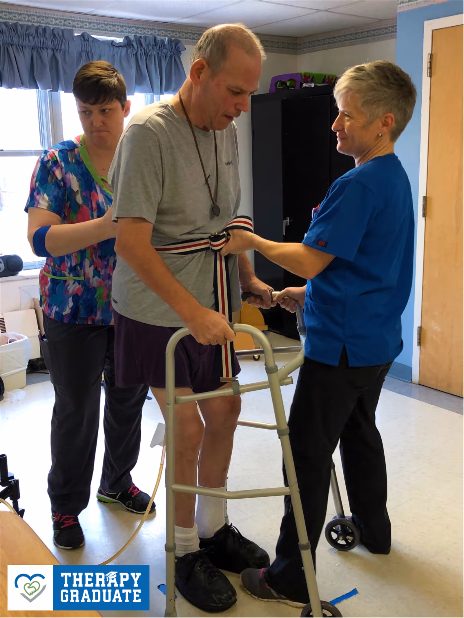 A man using a walker is assisted by two healthcare workers in a therapy room. One worker is helping to secure a belt around the man's waist while the other supports him from behind. The room has a window with blue curtains and a wooden door. A logo in the bottom-left corner reads 'THERAPY GRADUATE' with a heart and hand icon.