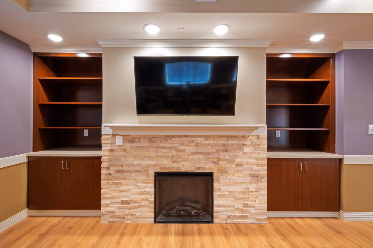 A cozy living room area featuring a stone fireplace with a mounted flat-screen TV above it. On either side of the fireplace are built-in wooden shelves and cabinets with a dark finish. The walls are painted in a combination of light beige and purple, and the floor is a light wood laminate.
