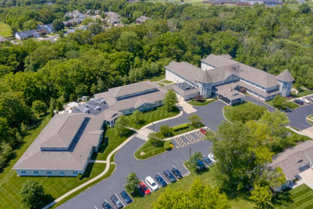 Aerial view of Addington Place of Shiloh senior living facility surrounded by green trees and lawns, showing multiple connected buildings with gray roofs, parking lots with cars, and paved walkways.