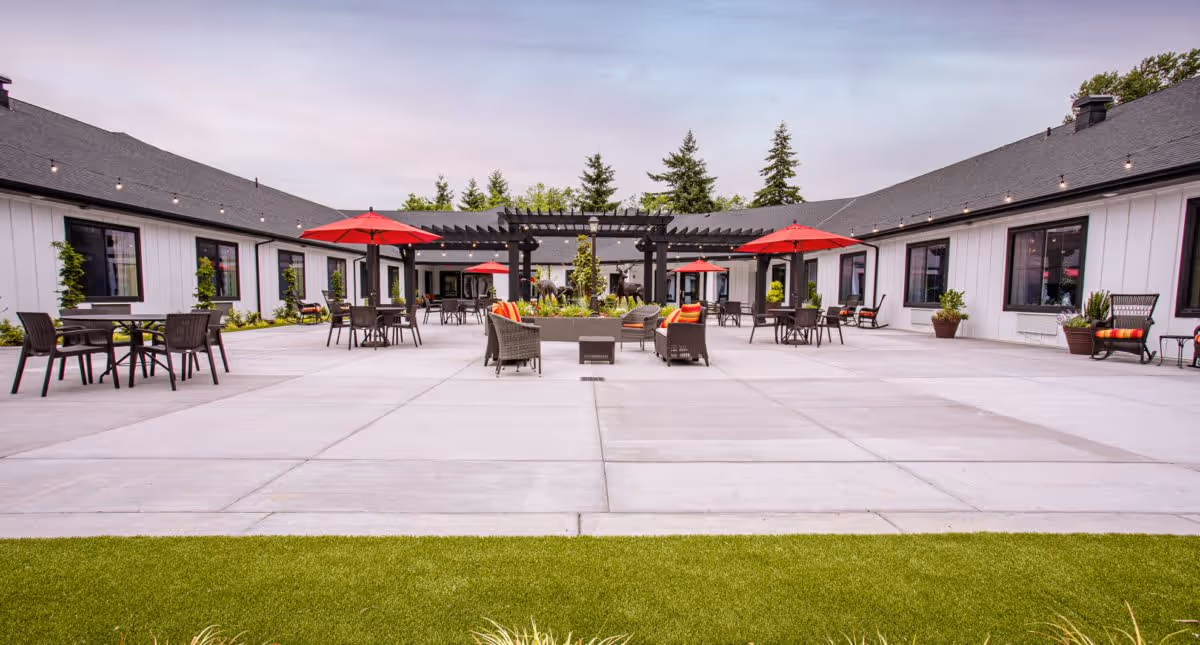 Outdoor courtyard area of Deer Ridge Memory Care featuring a spacious concrete patio with multiple seating arrangements including tables with red umbrellas and cushioned chairs. The courtyard is surrounded by a single-story building with white siding and black-framed windows. There is a pergola structure in the center with plants and decorative lighting strung overhead.
