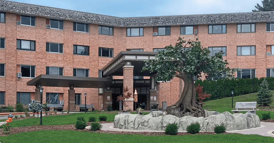 Exterior view of Schmitt Woodland Hills Retirement Community showing a multi-story brick building with numerous windows. In front of the building is a landscaped area with green grass, small bushes, a large artificial tree surrounded by rocks, benches, and a covered entrance.