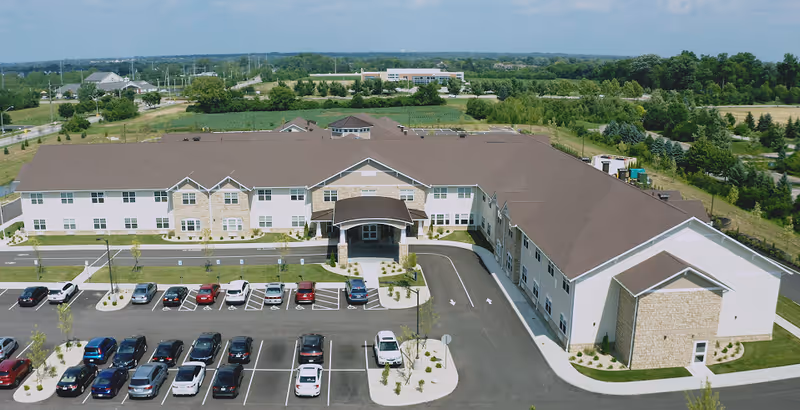 Aerial view of Norwich Springs Health Campus showing a large, two-story building with a brown roof and light-colored walls. The building has a covered entrance with a driveway and a parking lot filled with cars in front. Surrounding the building are landscaped areas with small trees and shrubs, and beyond the campus are open green fields and other buildings in the distance under a partly cloudy sky.