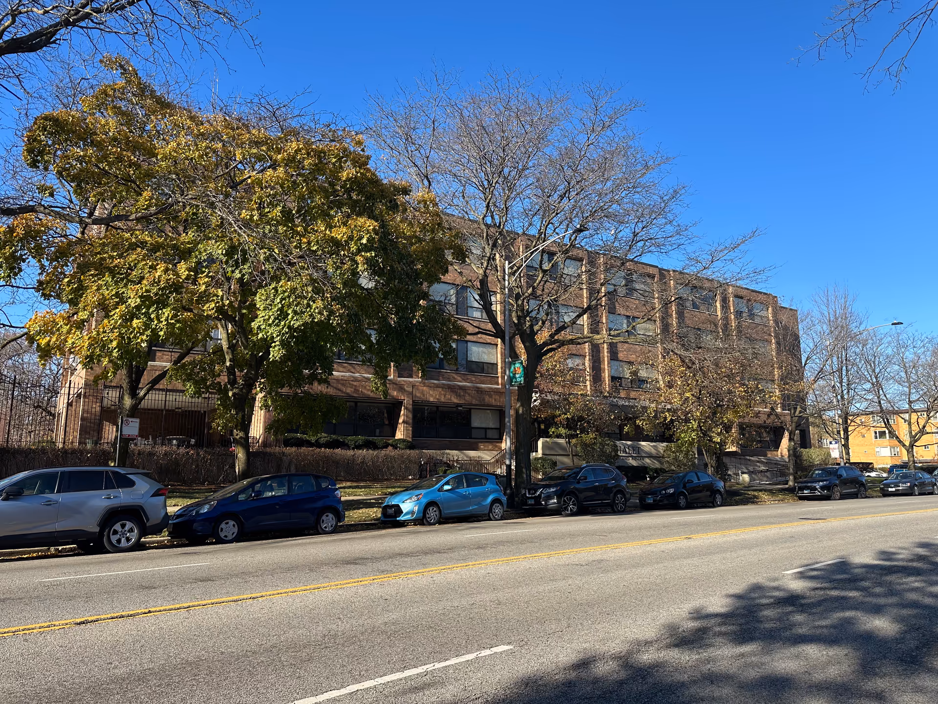 Exterior view of a multi-story brick building with several windows, partially obscured by trees with autumn foliage. Several cars are parked along the street in front of the building under a clear blue sky.