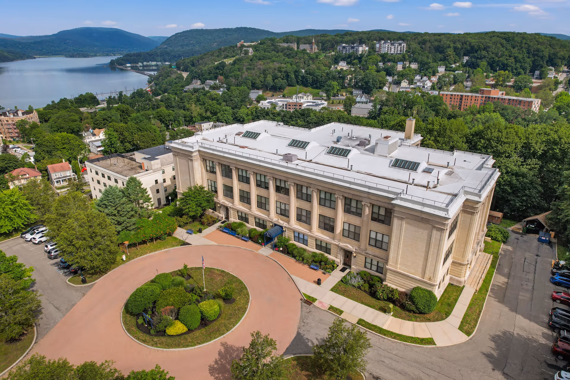 Aerial view of a large, classical-style senior living community building with a circular driveway and landscaped greenery in front. The building is surrounded by trees and other residential buildings, with a river and hills visible in the background under a partly cloudy sky.
