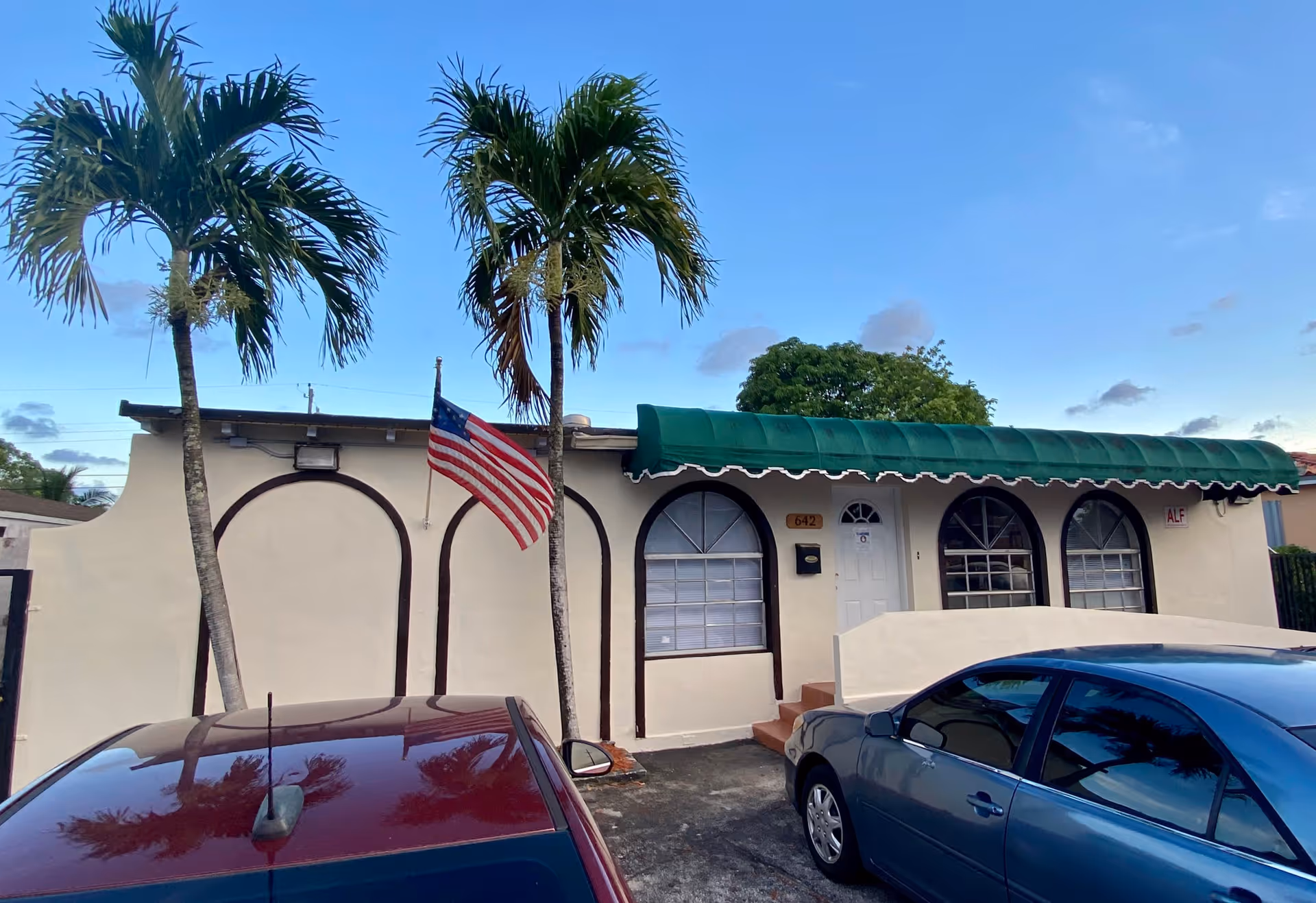 Exterior view of a single-story building with a green awning over the entrance, two palm trees in front, an American flag mounted on the wall, and two parked cars in the driveway under a clear blue sky.