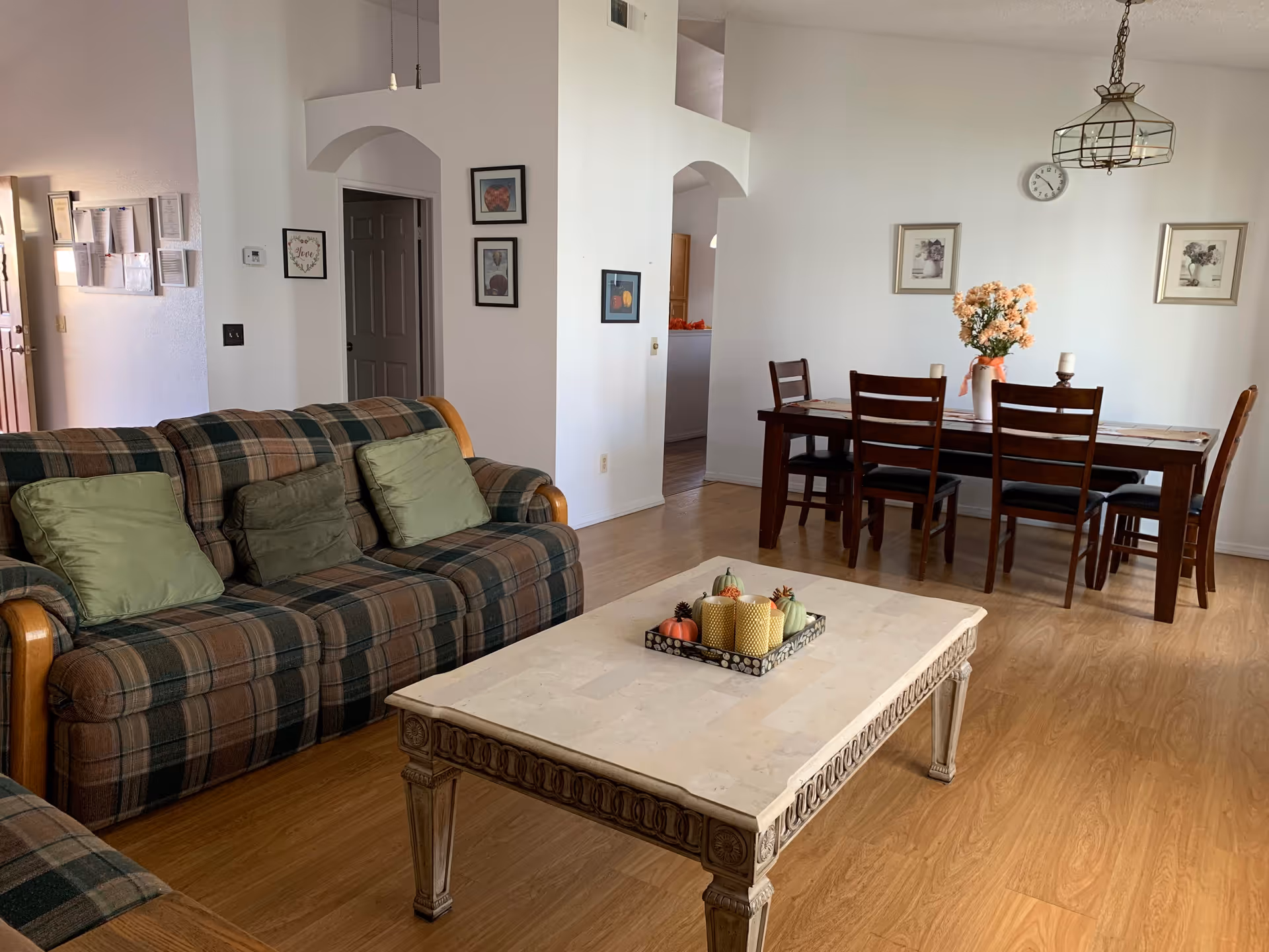 A cozy living and dining area with a plaid sofa adorned with green cushions, a decorative coffee table with candles and small pumpkins, and a wooden dining table with six chairs. The walls are white with framed artwork, and there is a hanging light fixture above the dining table. The floor is wooden.