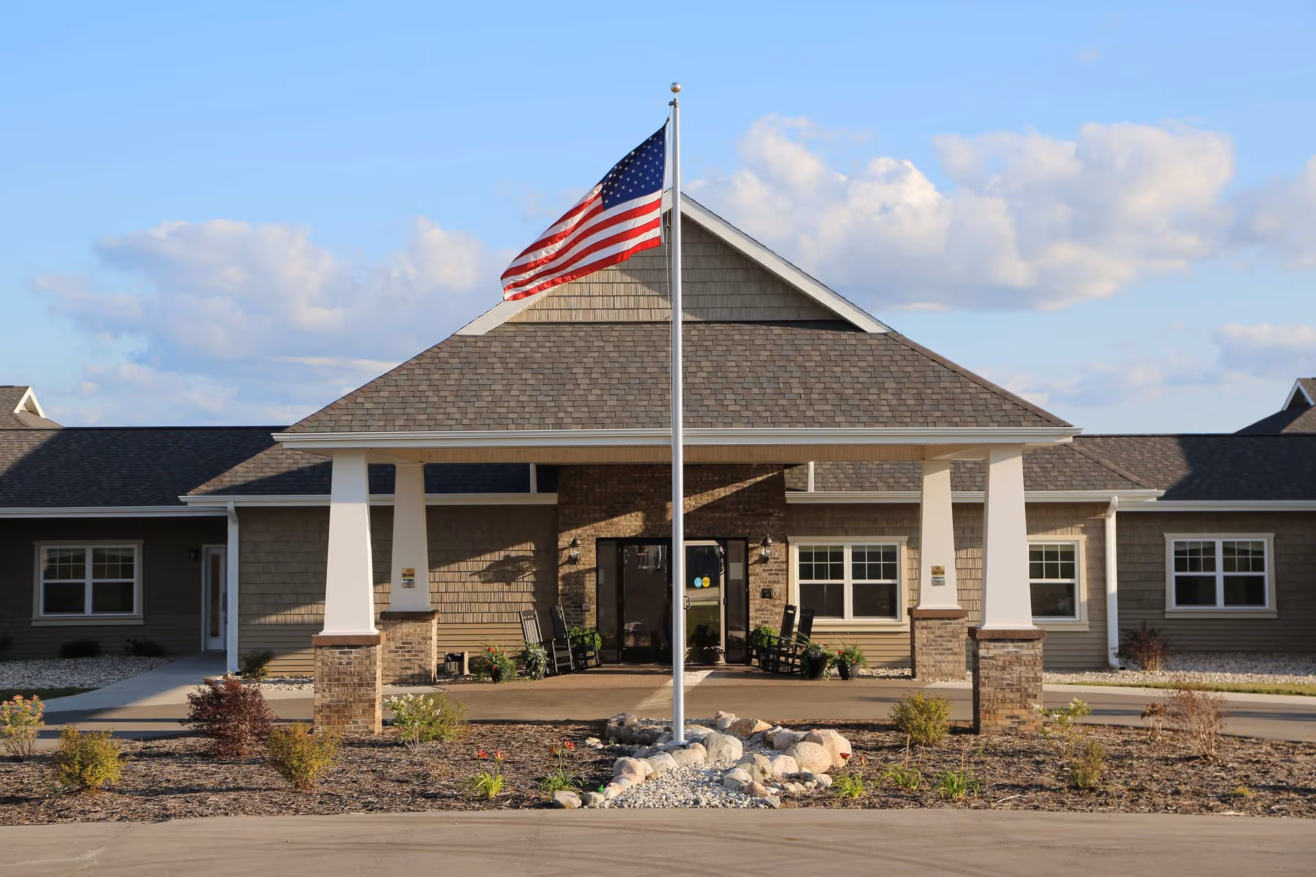 Front exterior view of The Brook of Cedar Springs facility with a covered entrance supported by white columns, an American flag on a flagpole in the center, and landscaped plants and rocks in front.