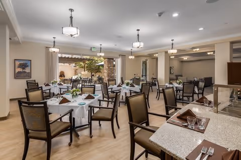 A spacious dining room with multiple round tables covered with white tablecloths, each set with brown napkins, plates, and glassware. The room features wooden chairs with cushioned seats, modern ceiling lights, and large windows that let in natural light. There are decorative plants and artwork on the walls, creating a welcoming atmosphere.