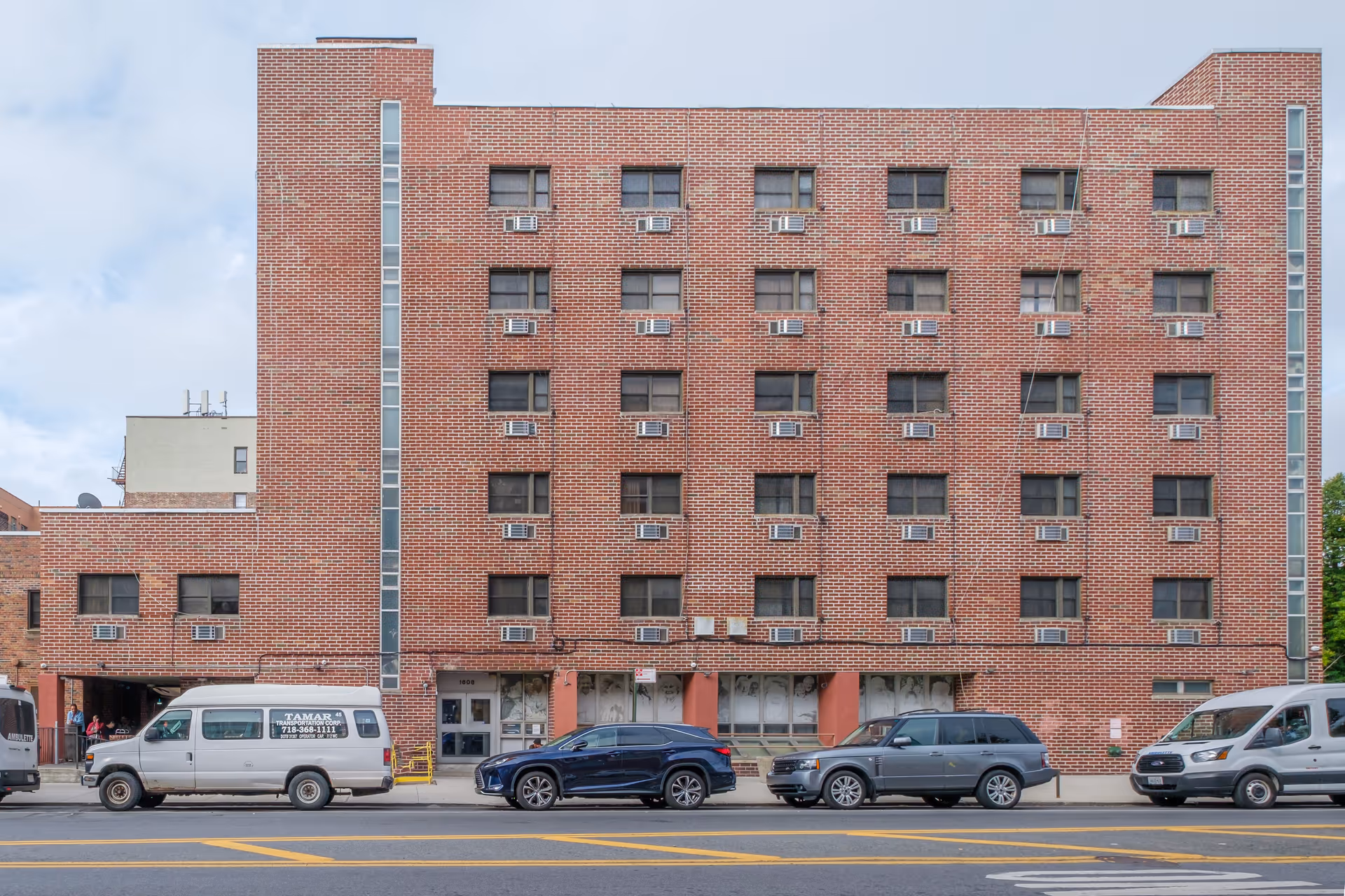 Front exterior view of a multi-story brick building with multiple windows, each equipped with window air conditioning units. Several vehicles are parked along the street in front of the building, including a white van with transportation company signage. The sky is partly cloudy.