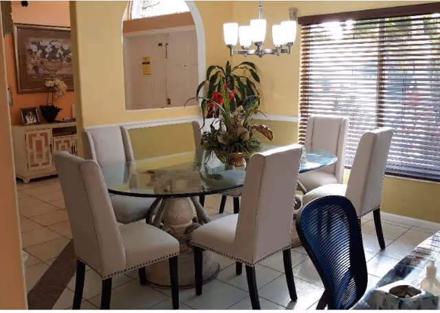 A dining area with a round glass table surrounded by six upholstered chairs. A decorative plant centerpiece is on the table. The room has yellow walls, a window with wooden blinds, and a chandelier hanging above the table. There is a blue office chair partially visible in the foreground and a sideboard with framed pictures and a flower arrangement in the background.