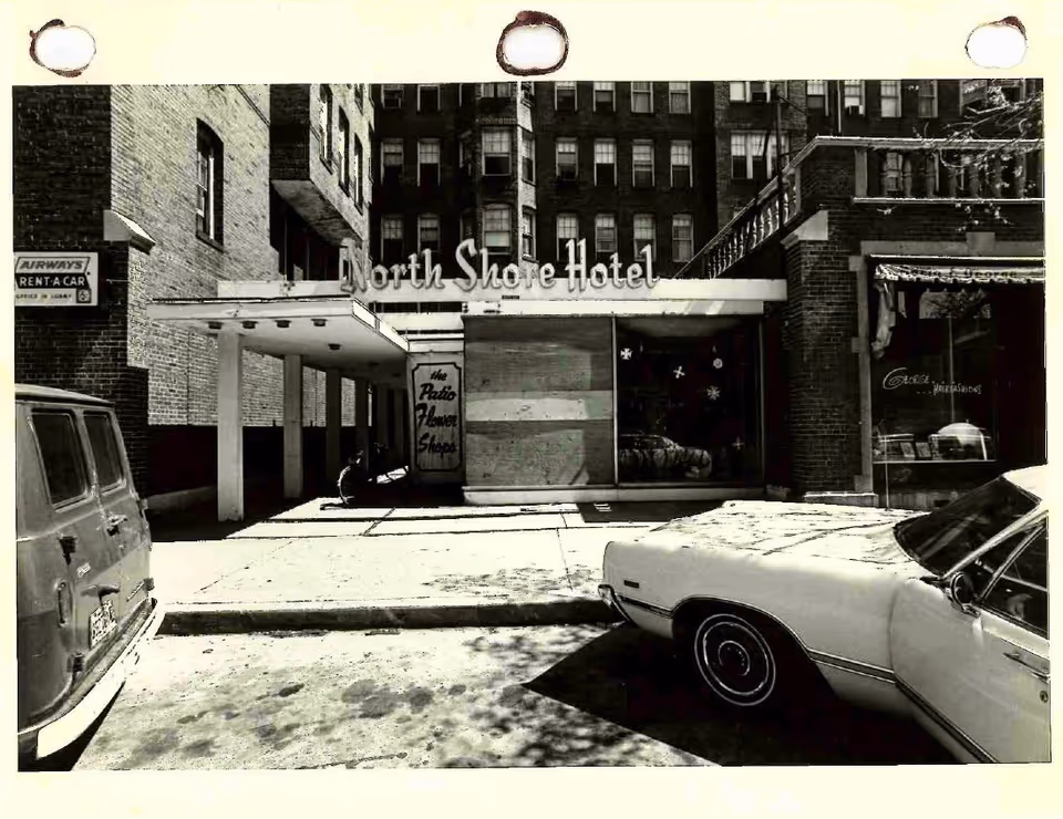Black and white photo of the exterior entrance of the North Shore Hotel with a covered walkway. Two cars are parked in front, one on the left and one on the right. There is a sign for Airways Rent-A-Car on the left side of the image and a sign for the Patio Flower Shop near the hotel entrance.