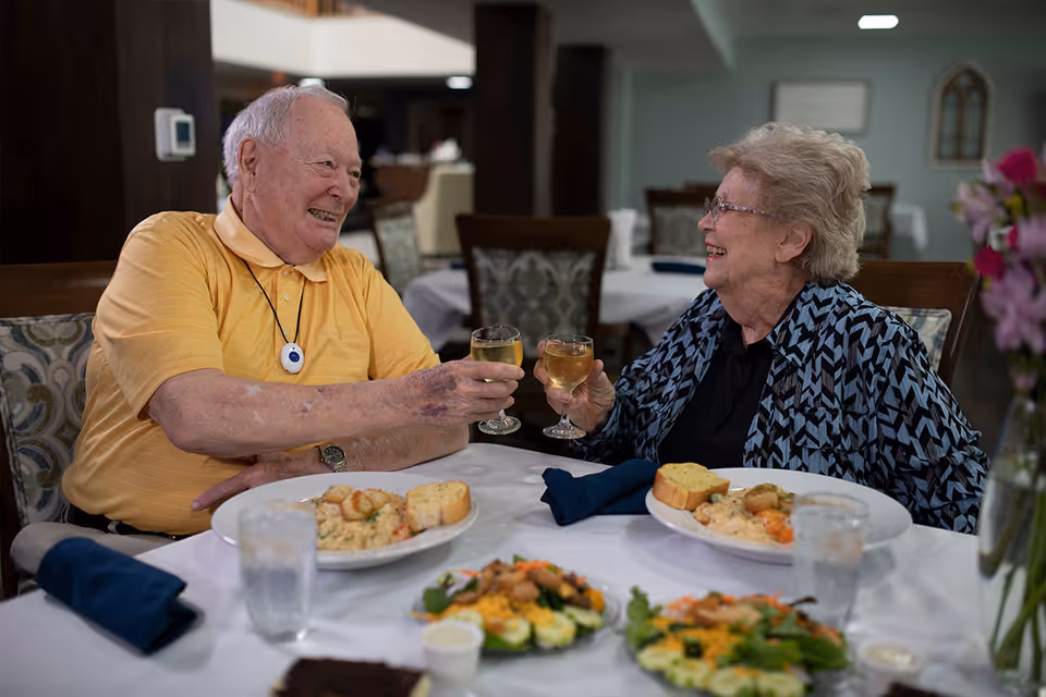 An elderly man and woman sitting at a dining table in a senior living facility, smiling and clinking glasses of white wine. Plates of food, including salad and bread, are on the table in front of them. The setting appears warm and inviting with a softly lit background.