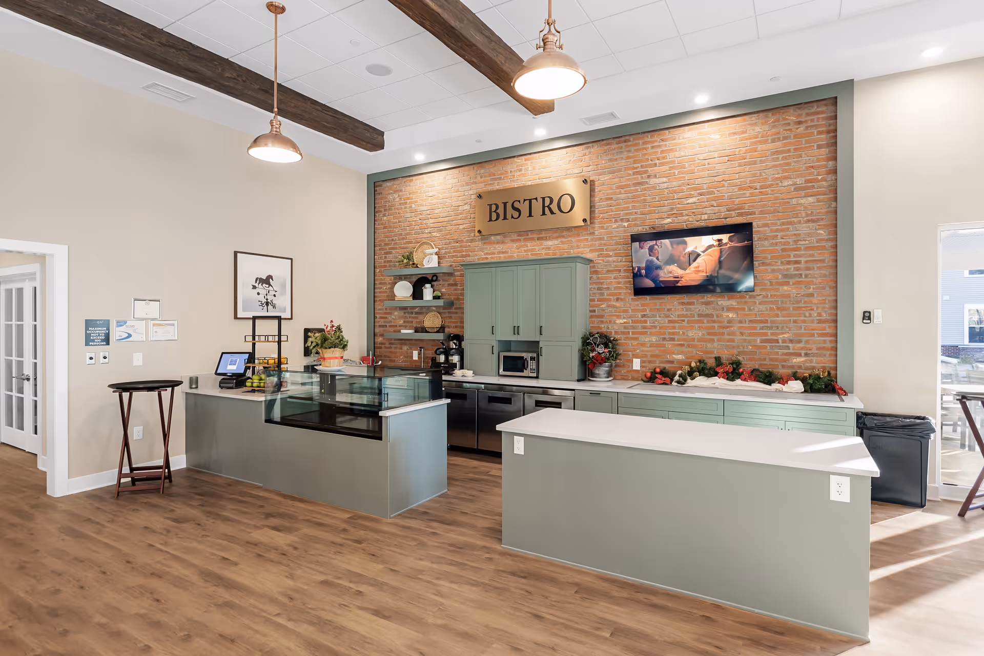 Interior view of a modern bistro area with light green cabinetry, a brick accent wall with a 'BISTRO' sign, a mounted TV, and wooden ceiling beams. The space includes a counter with a glass display case, a microwave, and decorative plants. The floor is wood, and there is natural light coming from windows on the right side.