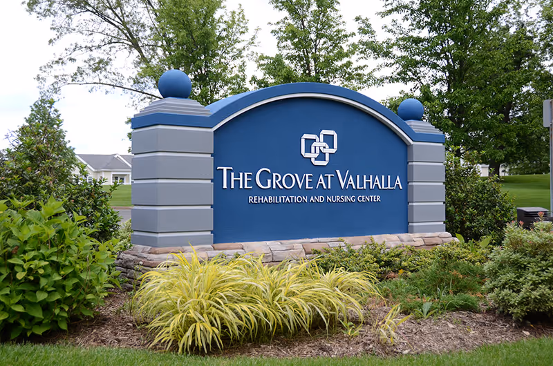 A large blue and gray stone sign for The Grove at Valhalla Rehabilitation and Nursing Center surrounded by green bushes and grass with trees in the background.