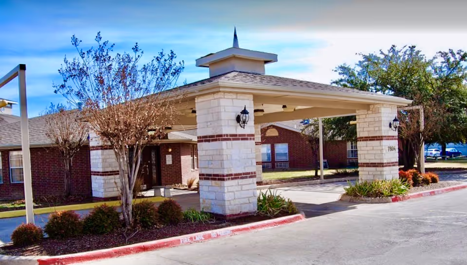 Entrance area of a nursing and rehabilitation facility with a covered drop-off zone supported by stone pillars. The building is made of red brick with white stone accents. There are small trees and shrubs planted around the entrance, and a clear blue sky is visible above.
