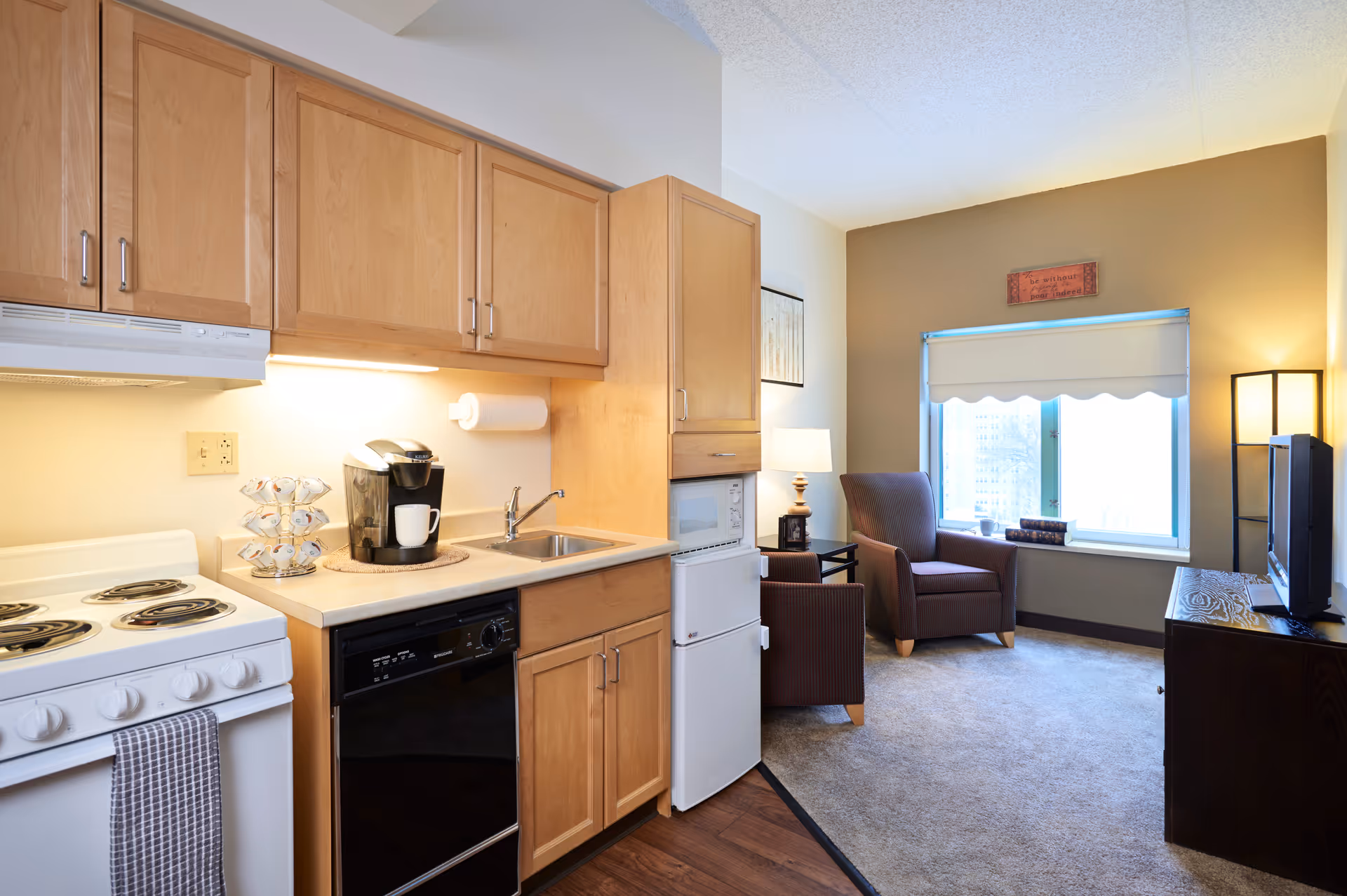 A small kitchen area with light wood cabinets, a white electric stove, a black dishwasher, a coffee maker, and a small sink. Adjacent to the kitchen is a cozy living space with two brown armchairs, a floor lamp, a TV on a wooden stand, and a window with a white scalloped blind letting in natural light.
