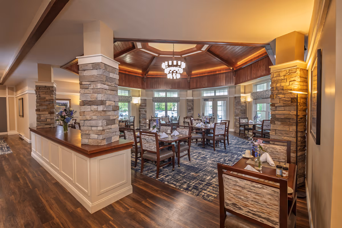 A spacious dining room with multiple wooden tables and chairs arranged neatly on a patterned carpet. The room features stone pillars, large windows letting in natural light, and a high wooden ceiling with a chandelier. The tables are set with white napkins and small flower vases, creating an inviting atmosphere.