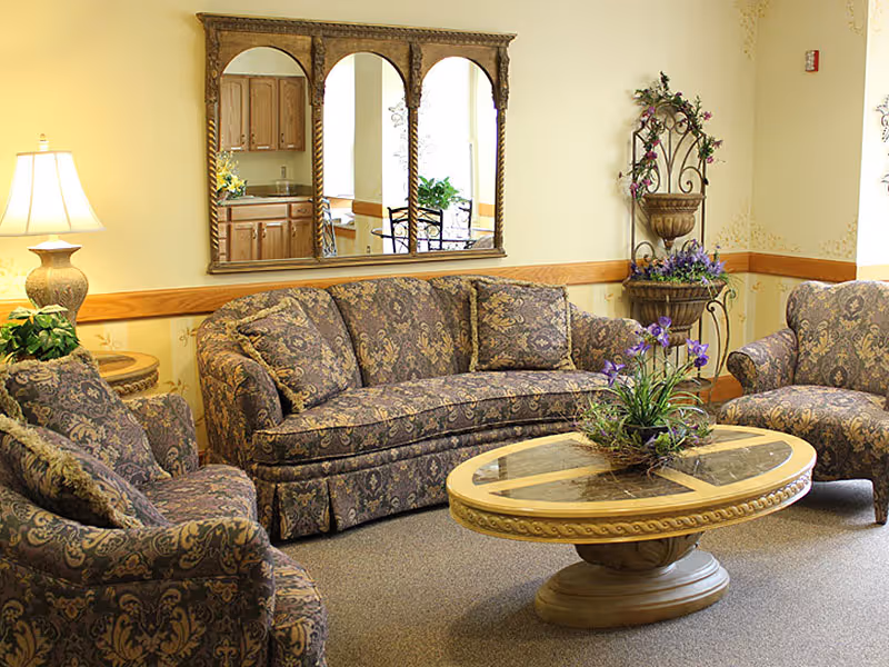 A cozy living room area in a retirement community featuring a patterned sofa and two matching armchairs arranged around an oval wooden coffee table with a glass top. A decorative plant centerpiece is on the table. A large ornate mirror with three arches hangs on the wall behind the sofa, reflecting a kitchen area. A table lamp and a decorative plant stand with purple flowers are also visible.