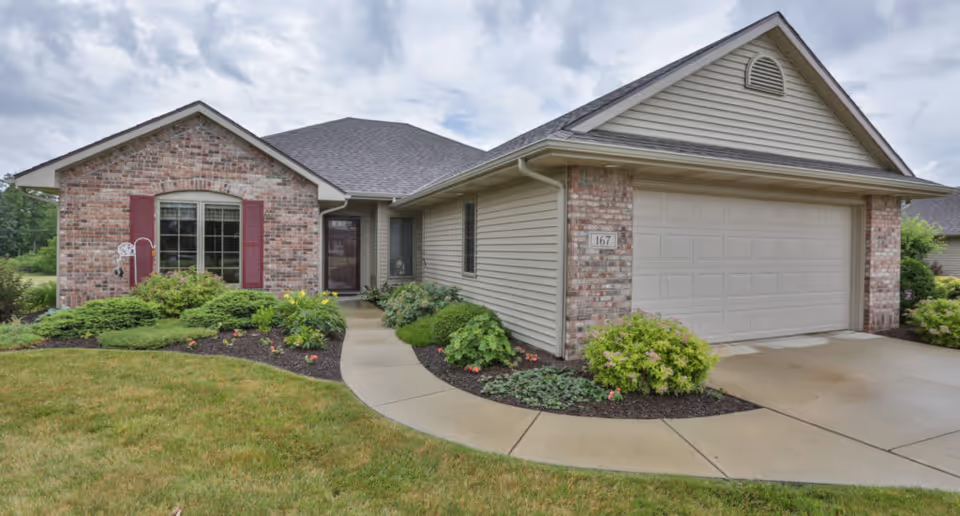 Exterior view of a single-story residential building with a brick and siding facade, a two-car garage, a curved concrete walkway leading to the front door, and landscaped bushes and grass under a cloudy sky.