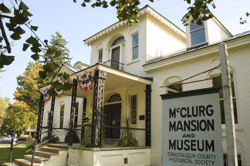 Exterior view of the McClurg Mansion and Museum, a historic white building with a porch decorated with red, white, and blue bunting. There are steps leading up to the entrance and a sign in front indicating it is part of the Chautauqua County Historical Society. Trees and greenery surround the building.