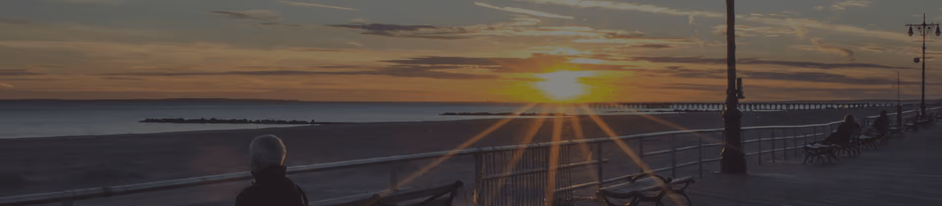 Sunset view over a beach with a pier extending into the water. A railing runs along the edge of a walkway where a few people are sitting on benches, enjoying the scenery.
