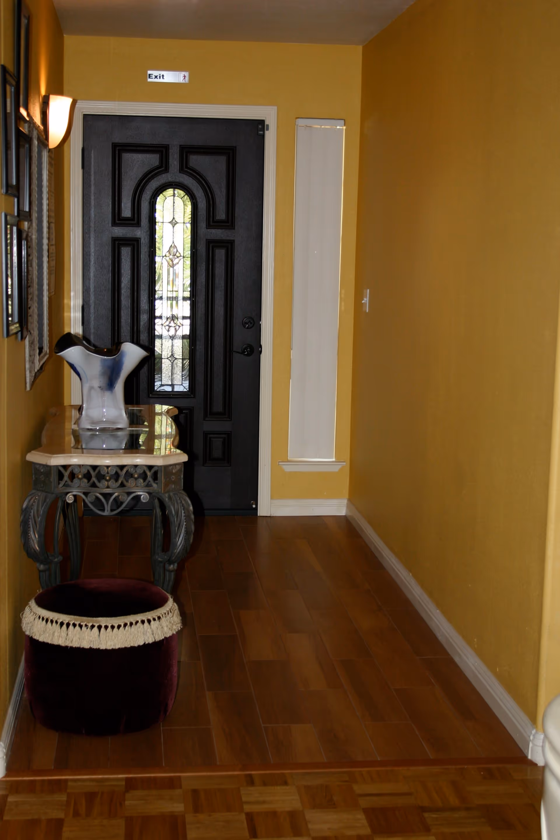 A narrow hallway with yellow walls and wooden flooring leading to a black door with decorative glass panels. On the left side, there is a small ornate table with a glass vase on top and a round purple ottoman with a fringe trim in front of it. A small vertical window with a white blind is on the right side of the door. A wall-mounted light fixture is visible on the left wall.
