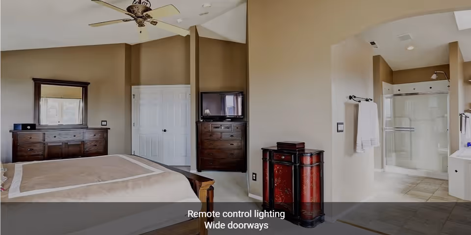A spacious bedroom with beige walls and a high ceiling featuring a ceiling fan. The room contains a large bed with a beige bedspread, a wooden dresser with a mirror, a chest of drawers with a TV on top, and a decorative red cabinet. To the right, there is an open doorway leading to a bathroom with a glass-enclosed shower and tiled floor. A towel rack with white towels is mounted on the wall near the bathroom entrance.