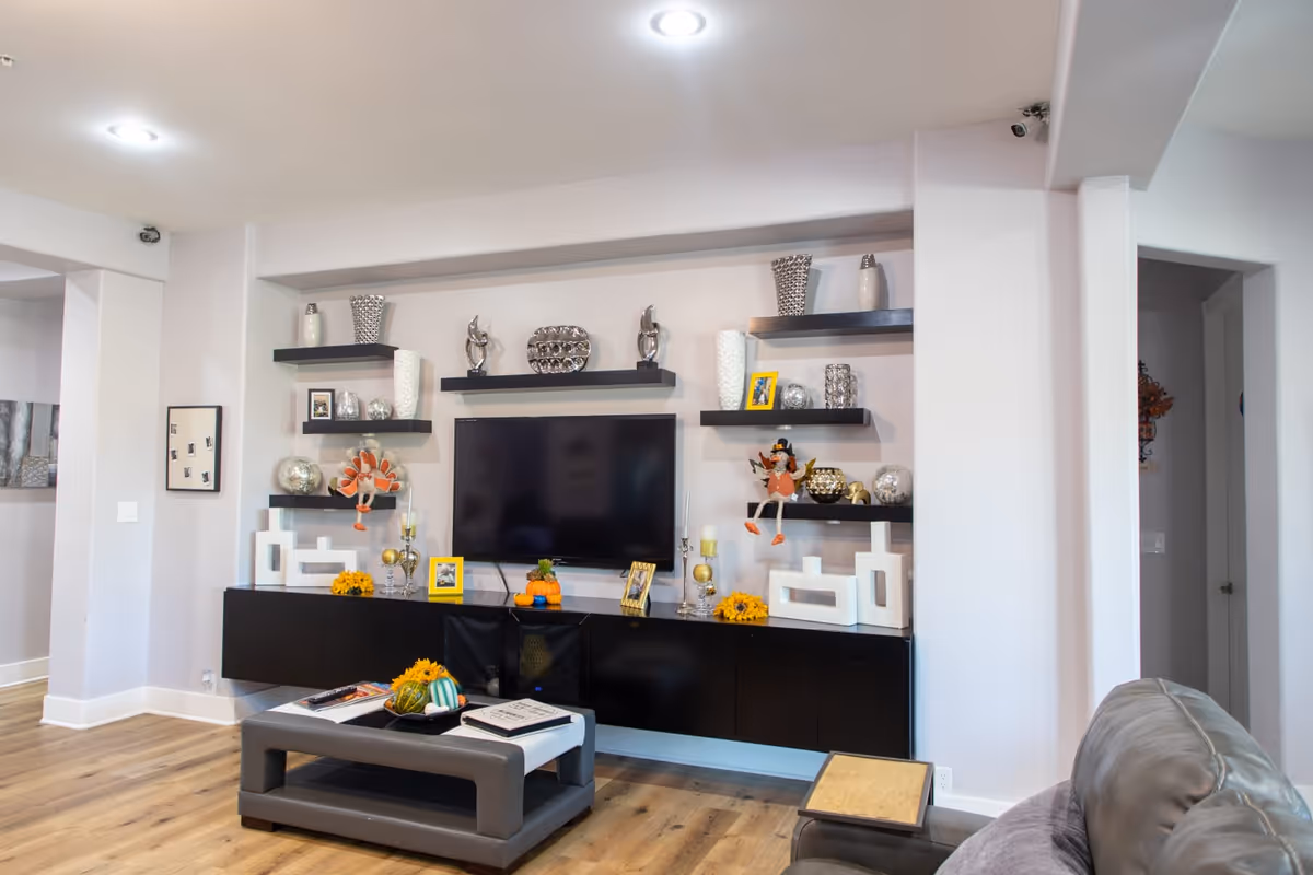 A modern living room with a wall-mounted flat-screen TV surrounded by black floating shelves displaying decorative items such as vases, sculptures, and framed photos. Below the TV is a long black cabinet with more decorations including small pumpkins and flowers. In front of the cabinet is a gray coffee table with books and a decorative pumpkin. The room has light-colored walls and wood flooring, with a gray leather chair partially visible on the right.
