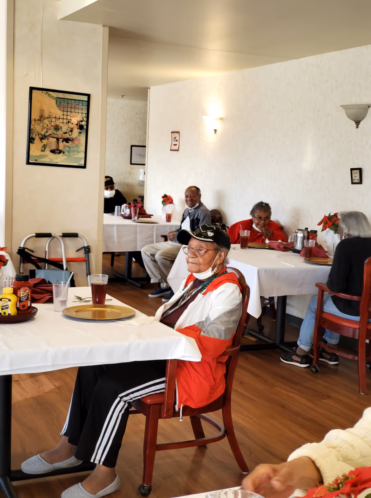Several elderly individuals seated at tables in a dining room with white tablecloths, red napkins, and drinks. The room has light-colored walls with framed pictures and wall sconces. One person in the foreground is wearing a red and white jacket and a black cap.