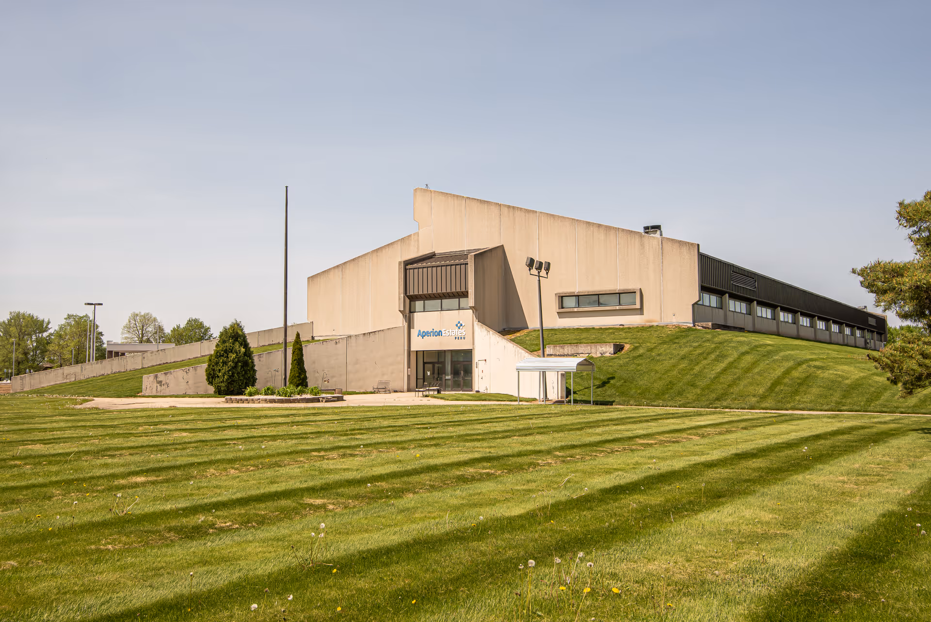 Exterior view of a large, modern building with a sloped grassy lawn in front. The building has a beige facade with a sign that reads Aperion Estates Peru above the entrance. The sky is clear and blue.