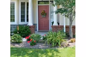 Front entrance of a house with a red door decorated with a wreath, flanked by white columns and brick walls. There are green shrubs, flowering plants, and a tree in the landscaped garden bed in front of the entrance.