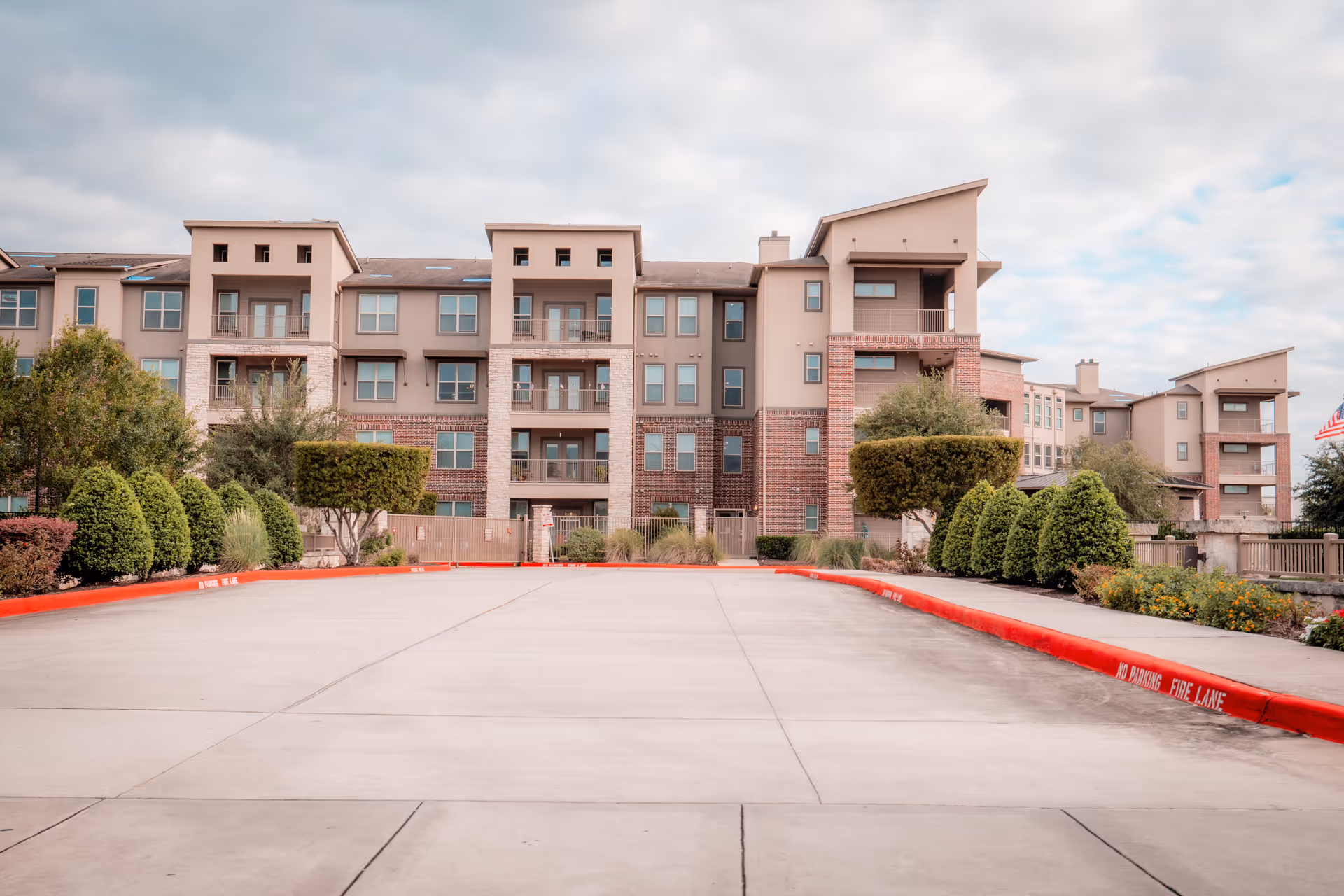 Exterior view of a multi-story residential building with balconies, surrounded by neatly trimmed bushes and trees under a cloudy sky. The foreground shows a wide concrete driveway with red curbs marked as a fire lane.