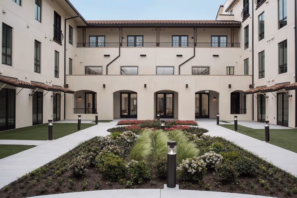 A landscaped courtyard area within a multi-story building featuring neatly trimmed bushes, ornamental grasses, and flower beds. The courtyard is surrounded by beige walls with multiple windows and doors, some with small balconies. Pathways and modern black bollard lights line the garden area.