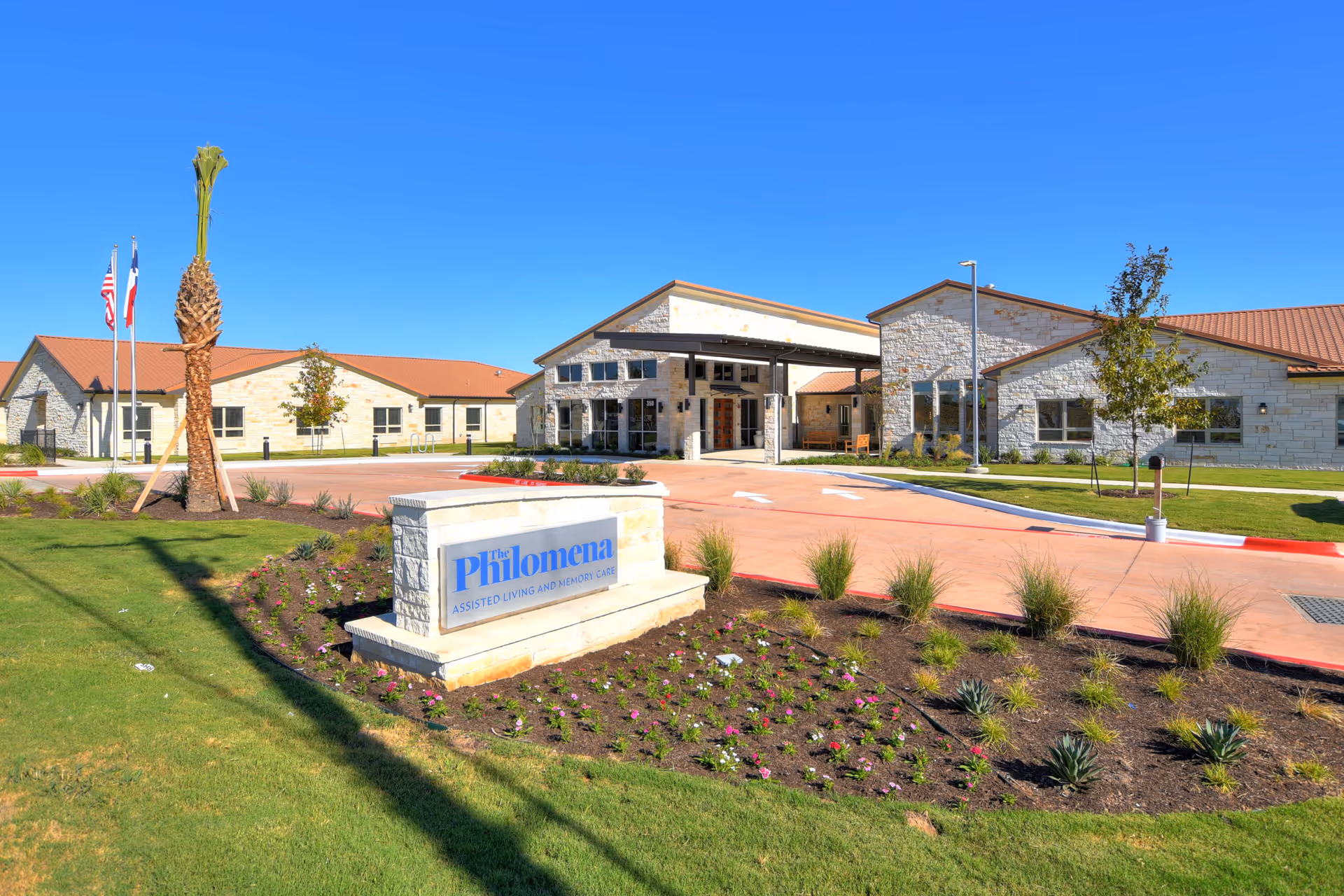 Exterior view of The Philomena assisted living and memory care facility with a stone sign in the foreground, landscaped garden beds, green lawn, and a clear blue sky. The building has light-colored stone walls and a red roof, with flags on poles near the entrance.