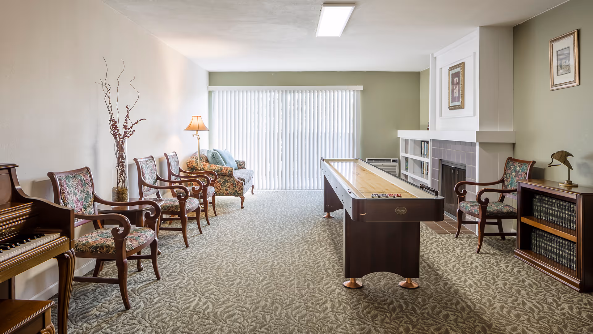 A well-lit living room with a shuffleboard table in the center, floral upholstered chairs lined up along the left wall, a floral sofa with cushions near a floor lamp, a fireplace with framed artwork above it, and a bookshelf with books and a small lamp. Vertical blinds cover a large window or sliding door at the far end of the room.